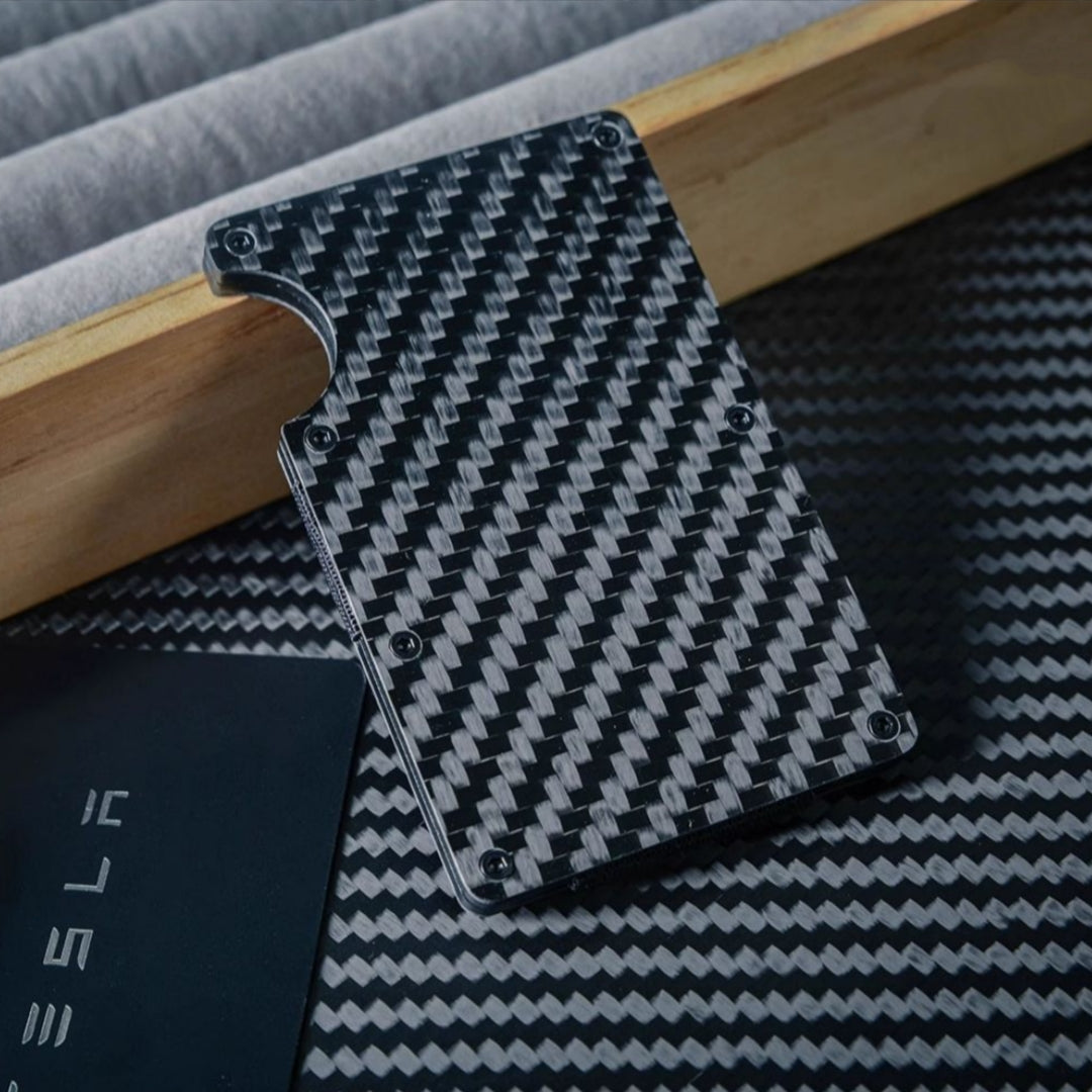 Close-up of a 3K carbon fiber card holder featuring a distinct woven pattern, secured with screw fasteners and a notched design for card access, placed near a Tesla-branded card on a carbon fiber-textured surface with wooden and soft lining accents.