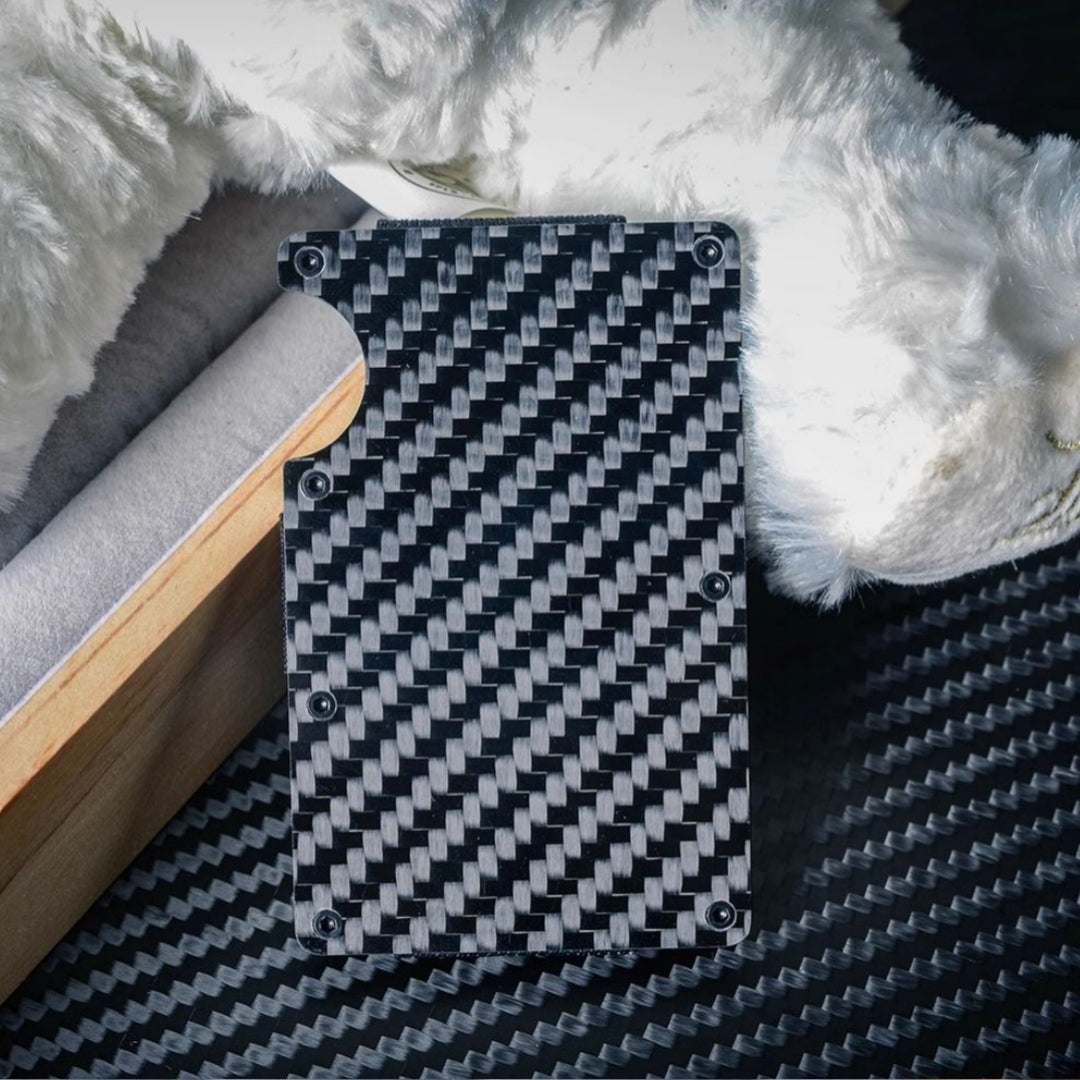 Close-up of a 3K carbon fiber card holder with a distinct woven pattern, secured by screw fasteners and featuring a notched design for easy card access, placed on a carbon fiber-textured surface with wooden, soft-lined, and plush accents in the background.