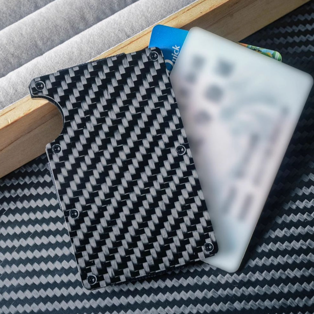 Close-up of a 3K carbon fiber card holder with a distinct woven pattern (secured by screw fasteners and featuring a notched design for easy card access), shown with inserted cards, placed on a carbon fiber-textured surface with wooden and soft-lined accents in the background.