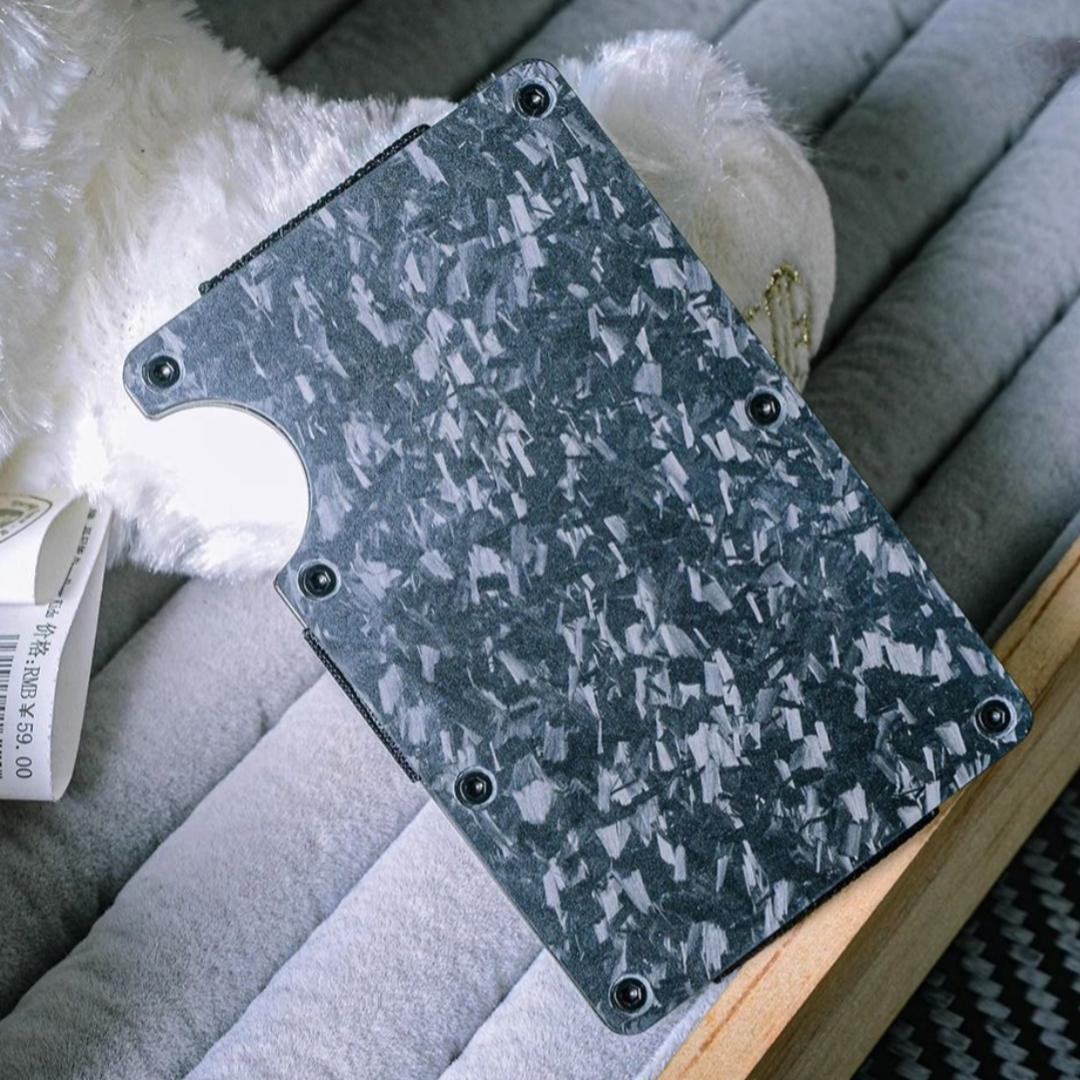 Close-up of a forged carbon fiber card holder with a distinct marbled texture, secured by screw fasteners and featuring a notched design for easy card access, placed on a soft, plush-lined surface with wooden accents in the background.