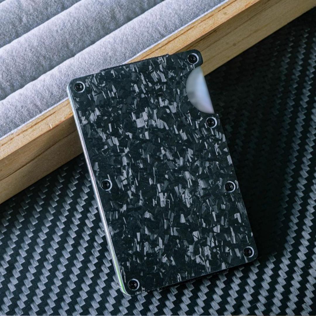 Close-up of a forged carbon fiber card holder with a distinct marbled texture, secured by screw fasteners and featuring a notched design for easy card access, placed on a carbon fiber-textured surface with wooden and soft-lined accents in the background.