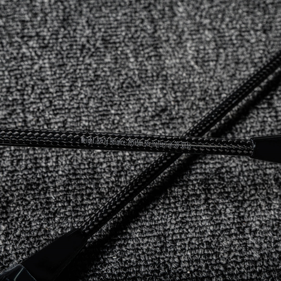 Close-up of two carbon fiber badminton racket shafts showcasing their distinct textured pattern and printed specifications, crossed on a gray textured surface.