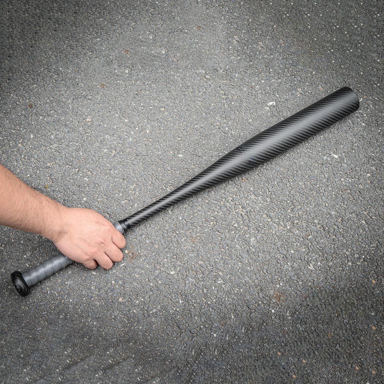 Hand holding a carbon fiber baseball bat with a distinct weave pattern, placed on an asphalt surface.