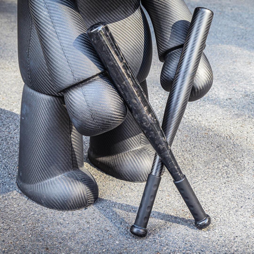 Two carbon fiber baseball bats with distinct weave patterns, crossed on an asphalt surface next to a large carbon fiber sculpture.