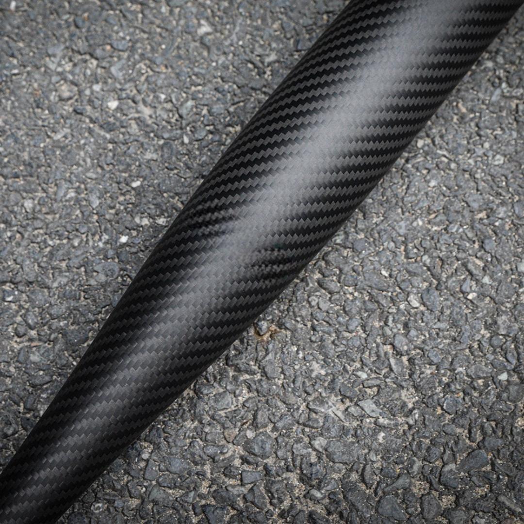 Close-up of a 3K carbon fiber baseball bat with a distinct weave pattern, placed on an asphalt surface.