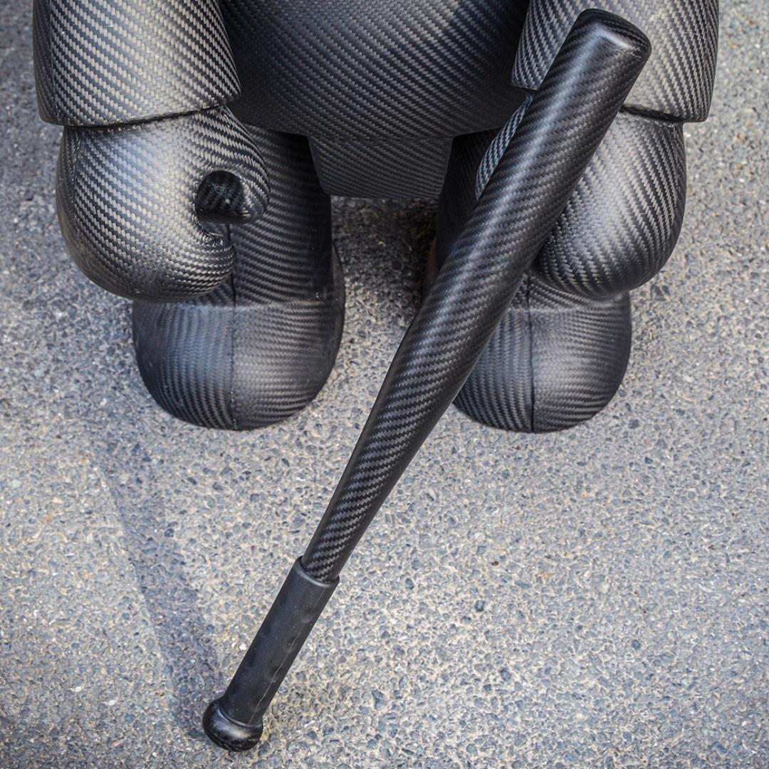 Close-up of a 3K carbon fiber baseball bat with a distinct weave pattern, positioned next to a large carbon fiber sculpture on an asphalt surface.