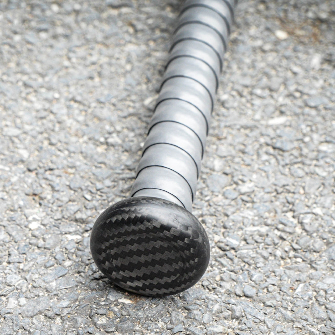 Close-up of the handle and end cap of a 3K carbon fiber baseball bat, showcasing the distinct carbon fiber weave pattern, placed on an asphalt surface.