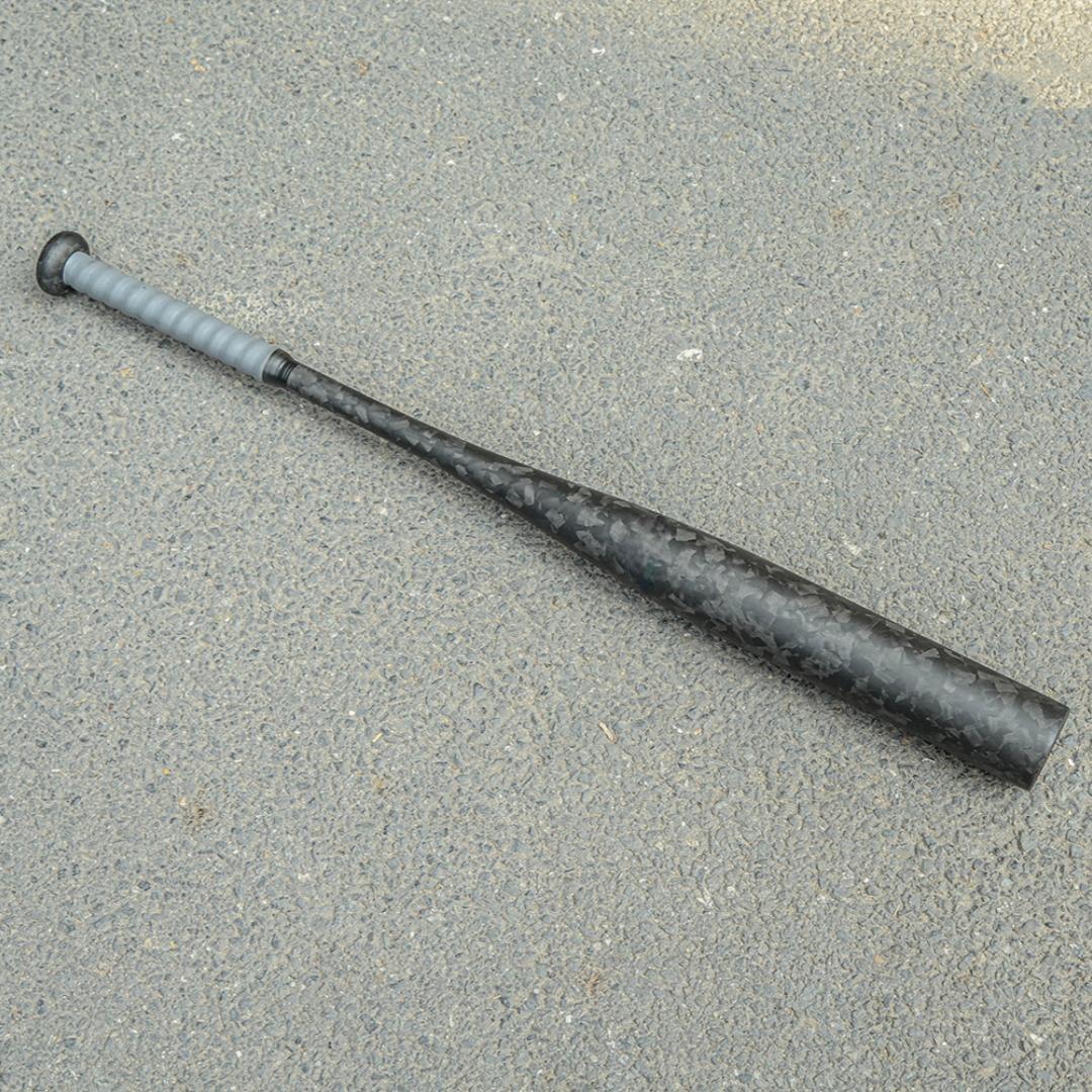 Forged carbon fiber baseball bat with a distinct chopped carbon fiber pattern and gray grip, laid on an asphalt surface.