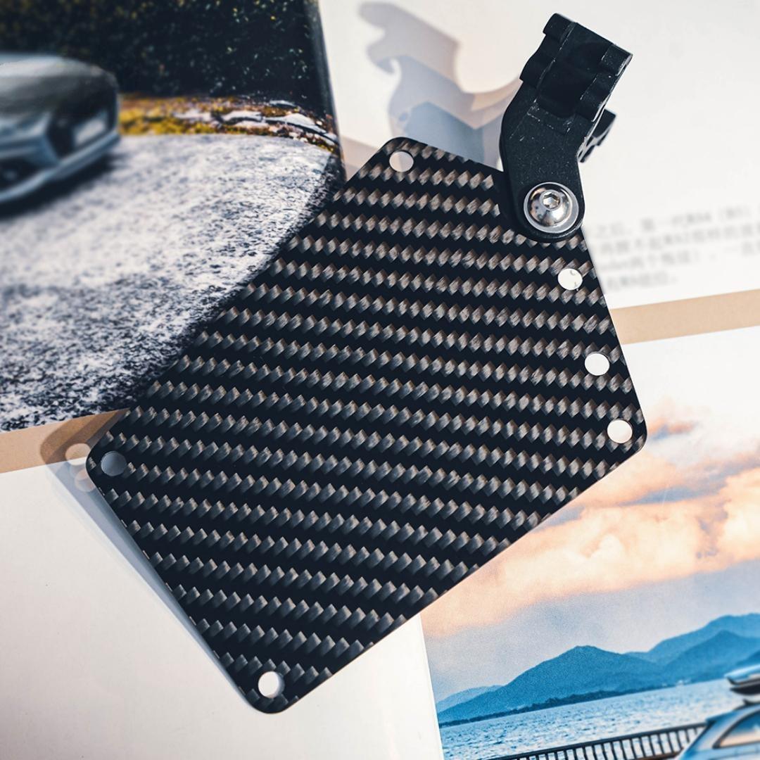 Close-up of a carbon fiber bicycle number plate with a distinct woven pattern, featuring a mounting clip, placed against a backdrop with automotive and landscape imagery.