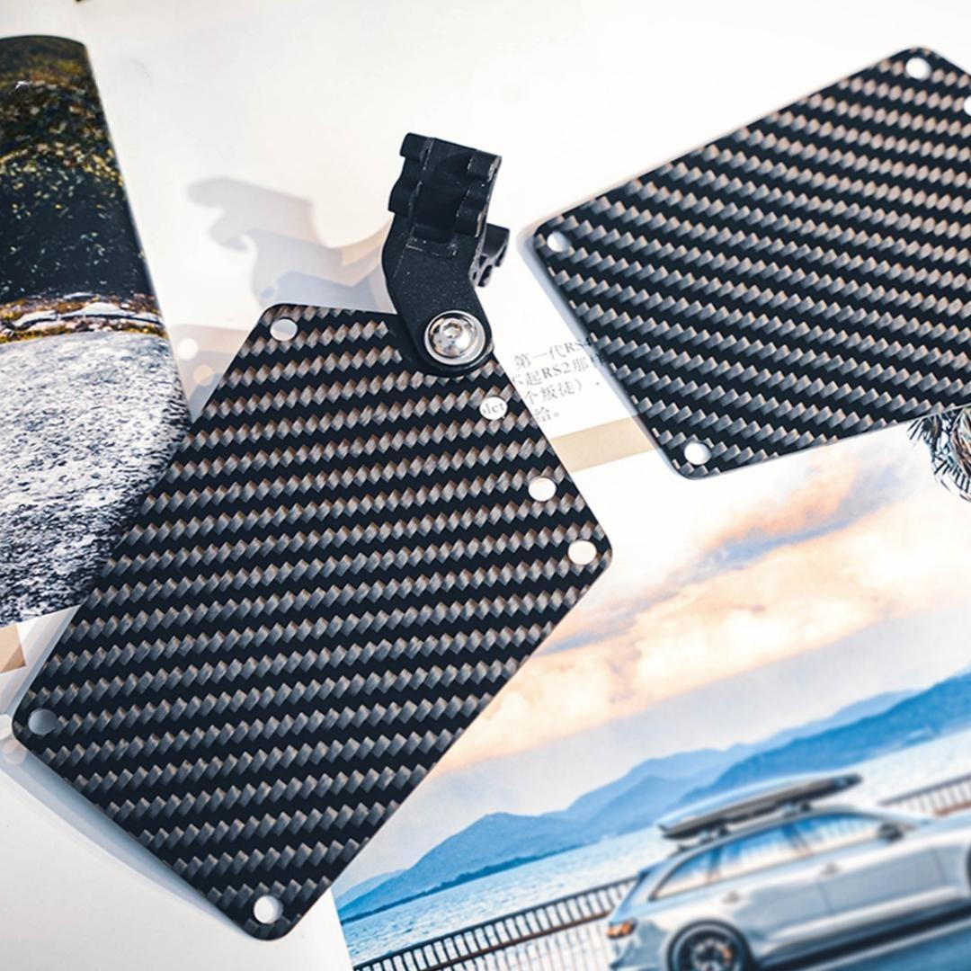 Close-up of two carbon fiber bicycle number plates with a distinct woven pattern, one featuring a mounting clip, displayed against a backdrop with automotive and landscape imagery.