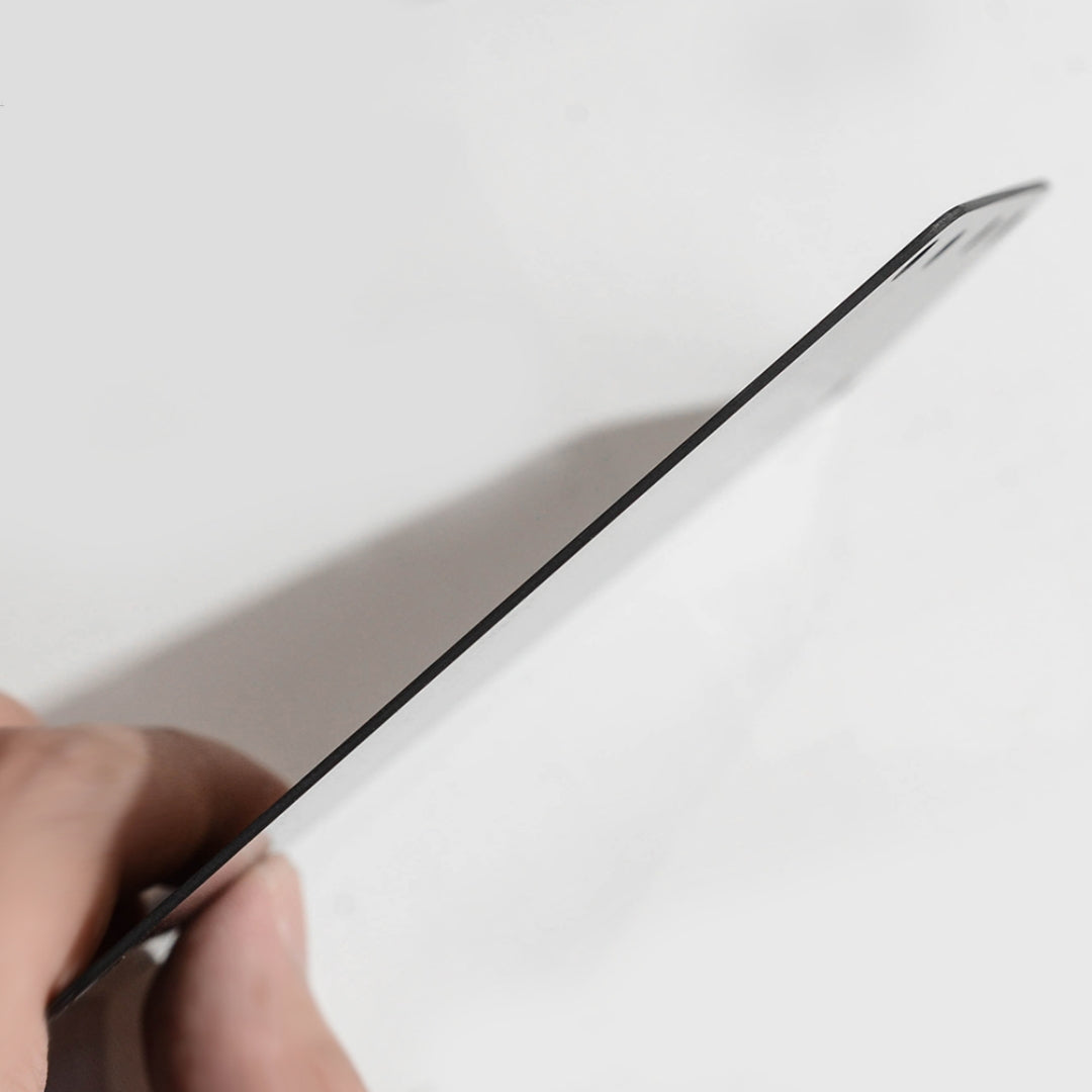 Close-up side view of a carbon fiber bicycle number plate, showcasing its thin profile, held by a hand against a white background.