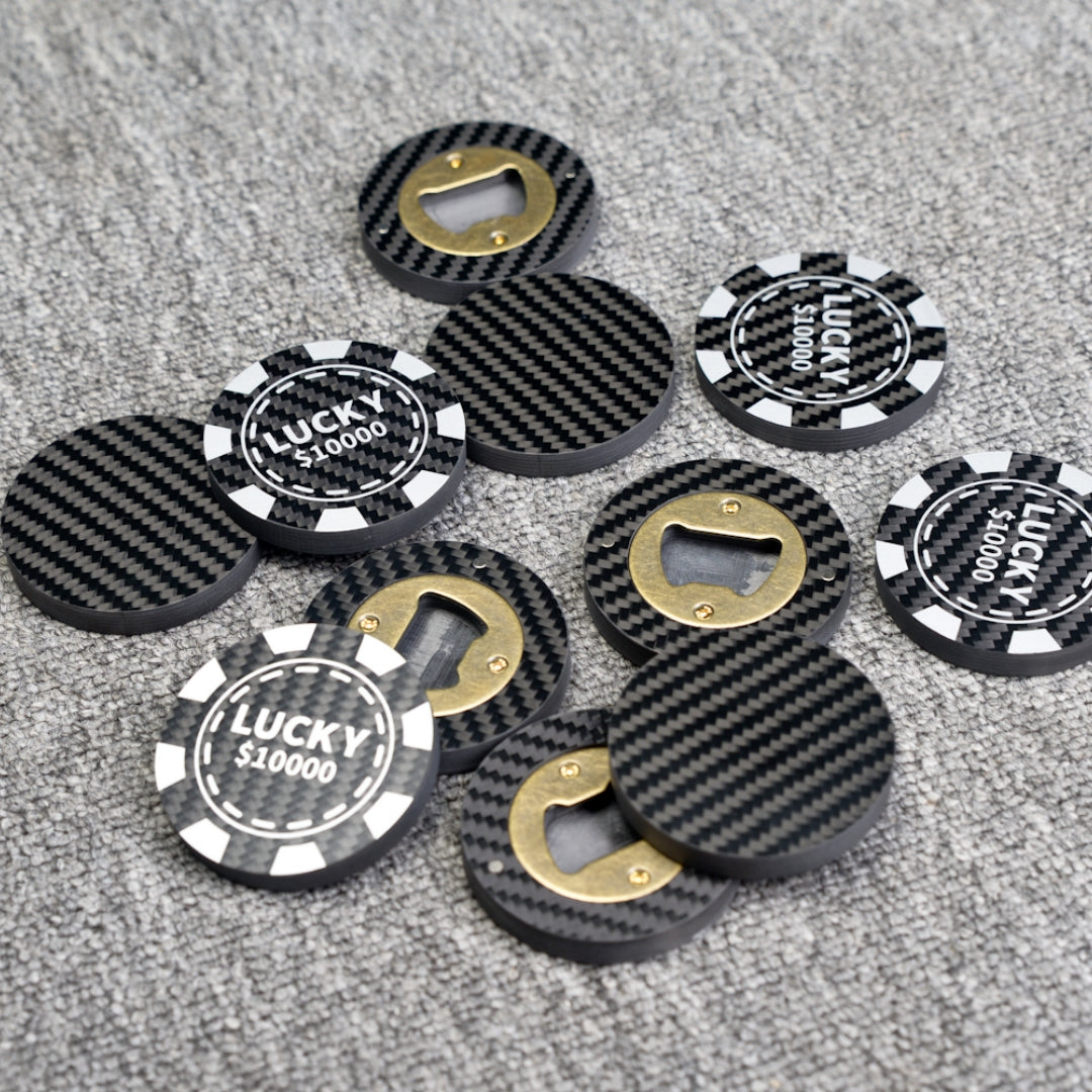 Close-up of multiple carbon fiber bottle openers with a poker chip design, featuring "LUCKY $10000" text and brass bottle opener mechanisms, scattered on a gray textured surface.
