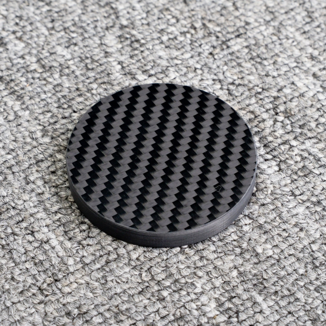 Close-up of a 3K carbon fiber bottle opener with a distinct woven pattern, showcasing its circular shape and glossy finish, placed on a gray textured surface.