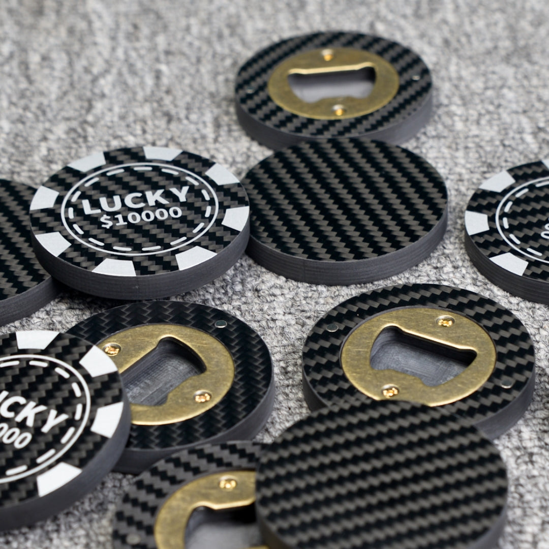 Close-up of multiple carbon fiber bottle openers, including poker chip-style ones with "LUCKY $10000" text and designs featuring brass bottle opener mechanisms, showcasing their distinct carbon fiber weave pattern while scattered on a gray textured surface.