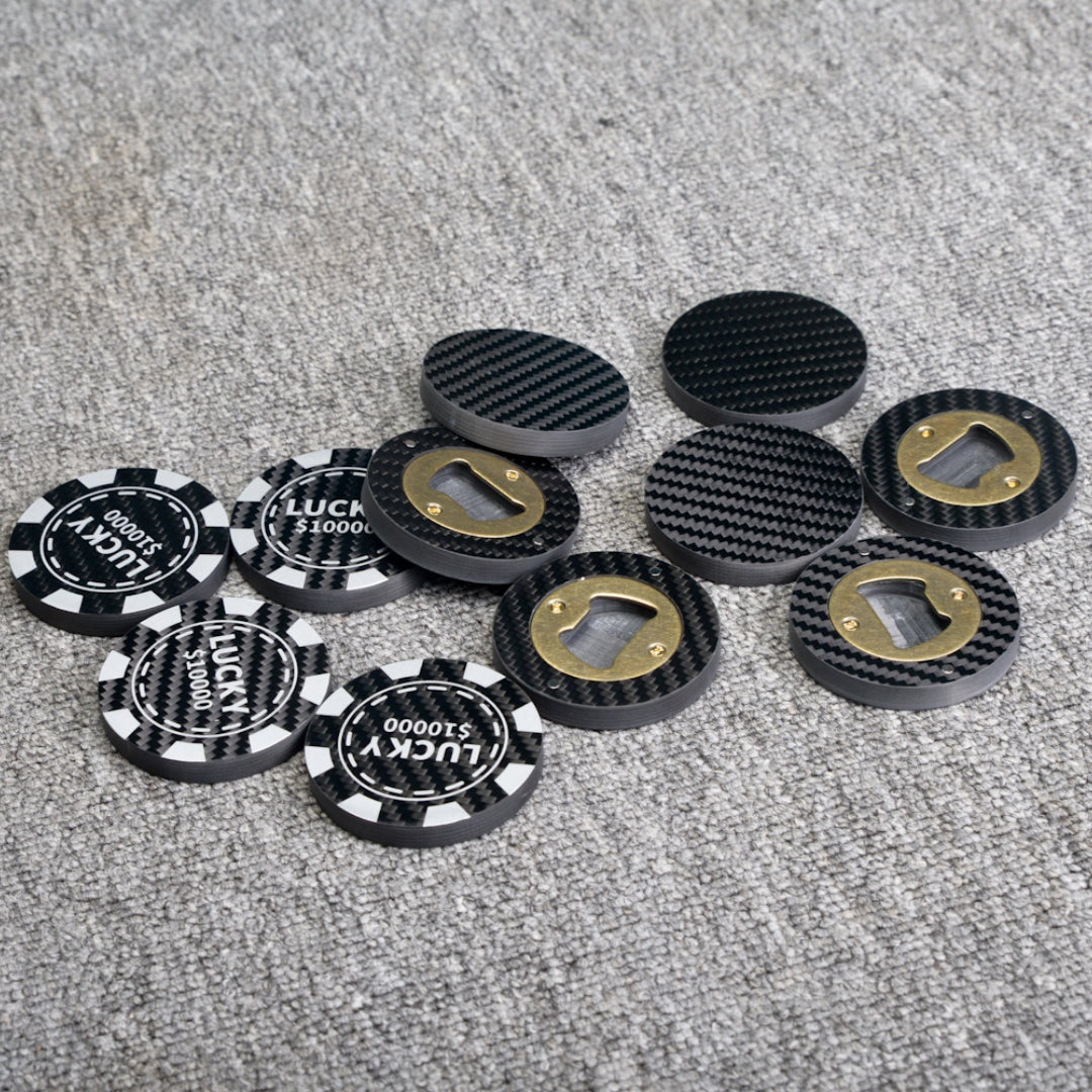 Close-up of multiple carbon fiber bottle openers, including poker chip-style ones with "LUCKY $10000" text and designs featuring brass bottle opener mechanisms, scattered on a gray textured surface.