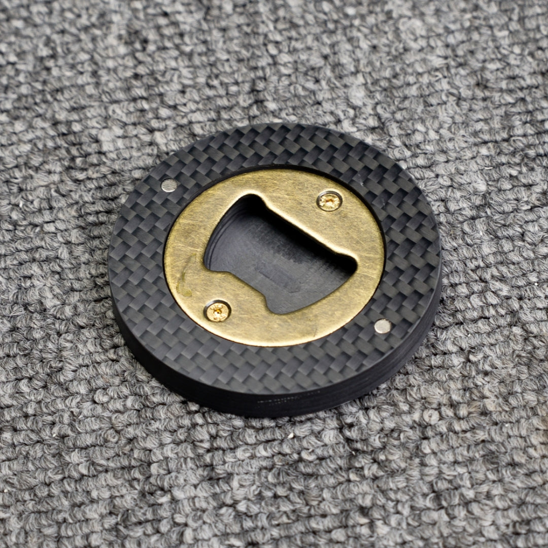 Close-up of a 3K carbon fiber bottle opener with a brass opener mechanism, showcasing its circular shape and distinct woven carbon fiber pattern, placed on a gray textured surface.