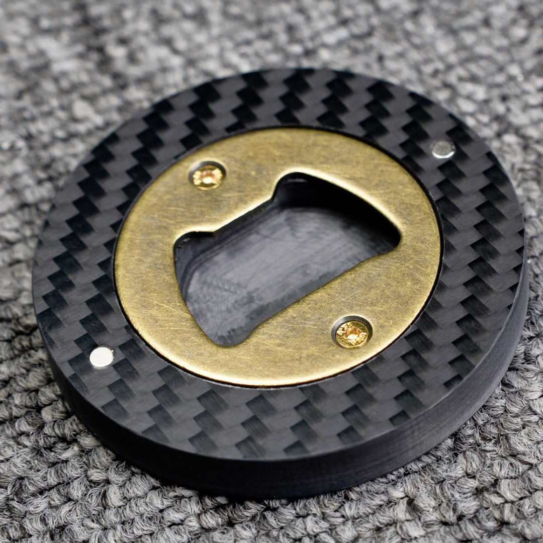 Close-up of a 3K carbon fiber bottle opener with a brass opener mechanism, showcasing its circular shape and distinct woven carbon fiber pattern, placed on a gray textured surface.