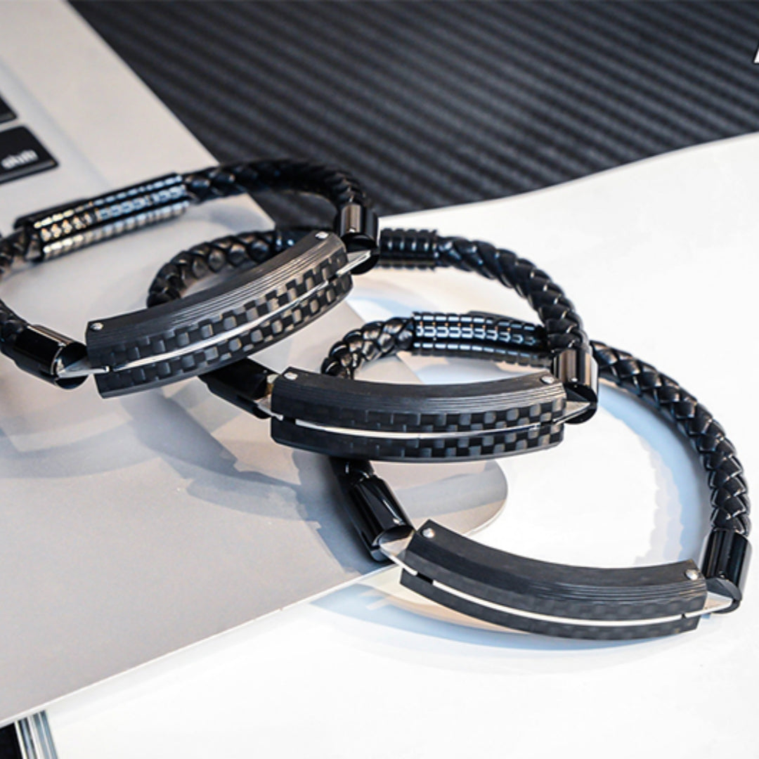 Close-up detail of several carbon fiber bracelets featuring black braided straps and metallic accents, placed on a light - colored surface with a laptop in the background.