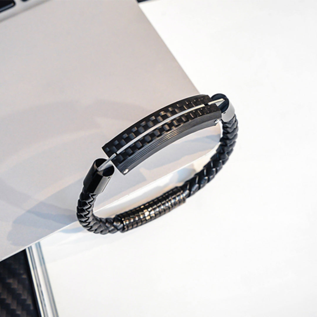 Close-up detail of a carbon fiber bracelet featuring a black braided strap and metallic accents, resting on a light-colored surface.