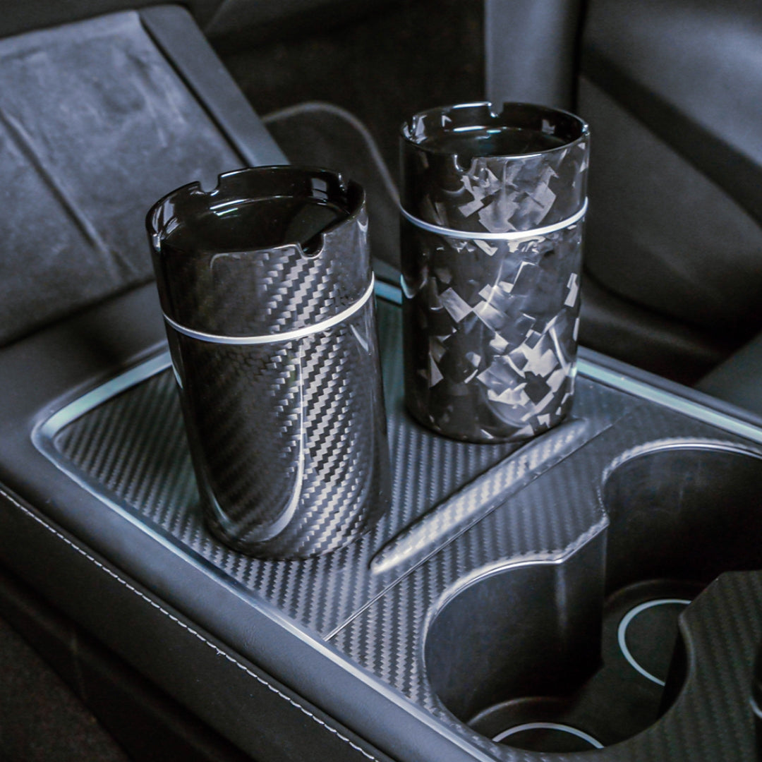 Close-up of two carbon fiber car ashtrays—one with a 3K carbon fiber pattern, the other with a forged carbon fiber pattern—placed on a carbon fiber-trimmed center console in a vehicle.
