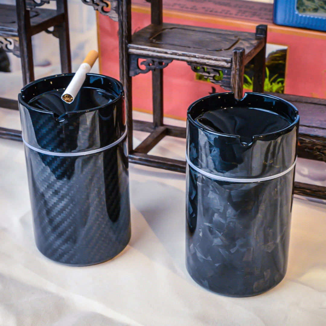 Close-up of two car ashtrays—one with a 3K carbon fiber pattern, the other with a forged carbon fiber pattern—with a cigarette in the left ashtray, displayed against traditional Chinese wooden decor.