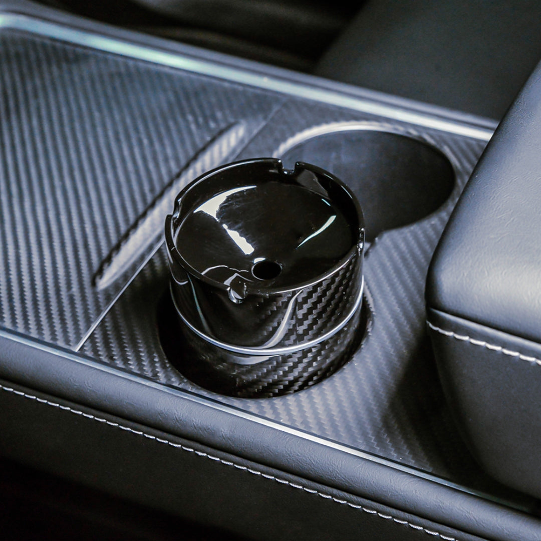 Close-up detail of a 3K carbon fiber car ashtray with a distinct woven pattern and glossy finish, placed in the cup holder of a car's center console featuring carbon fiber trim and leather upholstery.