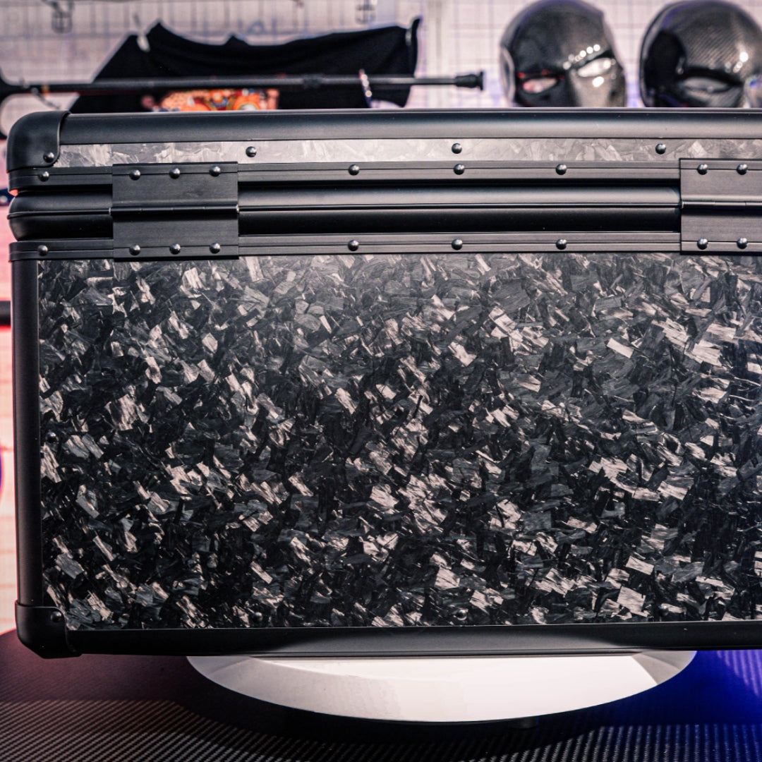Close-up detail of the back side of a large forged carbon fiber car storage trunk, showcasing the chopped carbon fiber pattern, metallic rivets, and structural details, with decorative masks visible in the background.