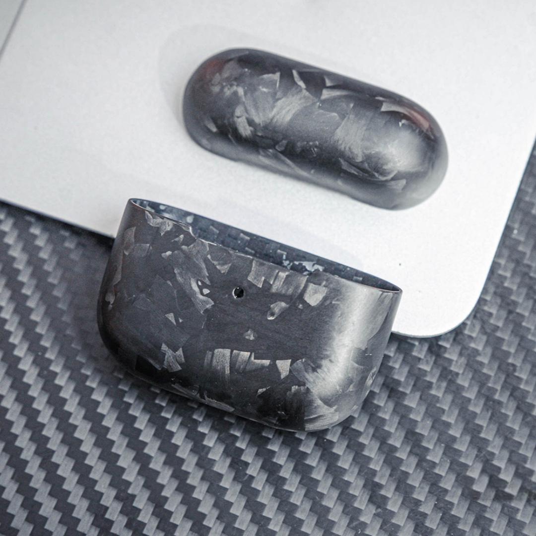 Close-up of forged carbon fiber AirPods cases showcasing the unique marbled texture, displayed on a surface with a carbon fiber pattern and a metallic background.