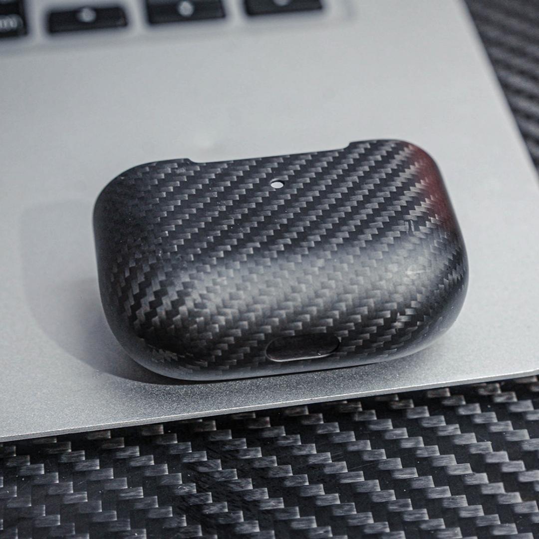 Close-up of the lower part of a 3K carbon fiber AirPods case showcasing the distinct weave pattern, placed on a metallic surface with a carbon fiber textured background.