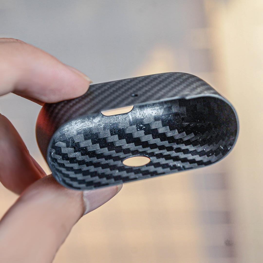 Close-up of the interior of the lower half of a 3K carbon fiber AirPods case, showcasing the distinct weave pattern, held in a hand against a soft background.