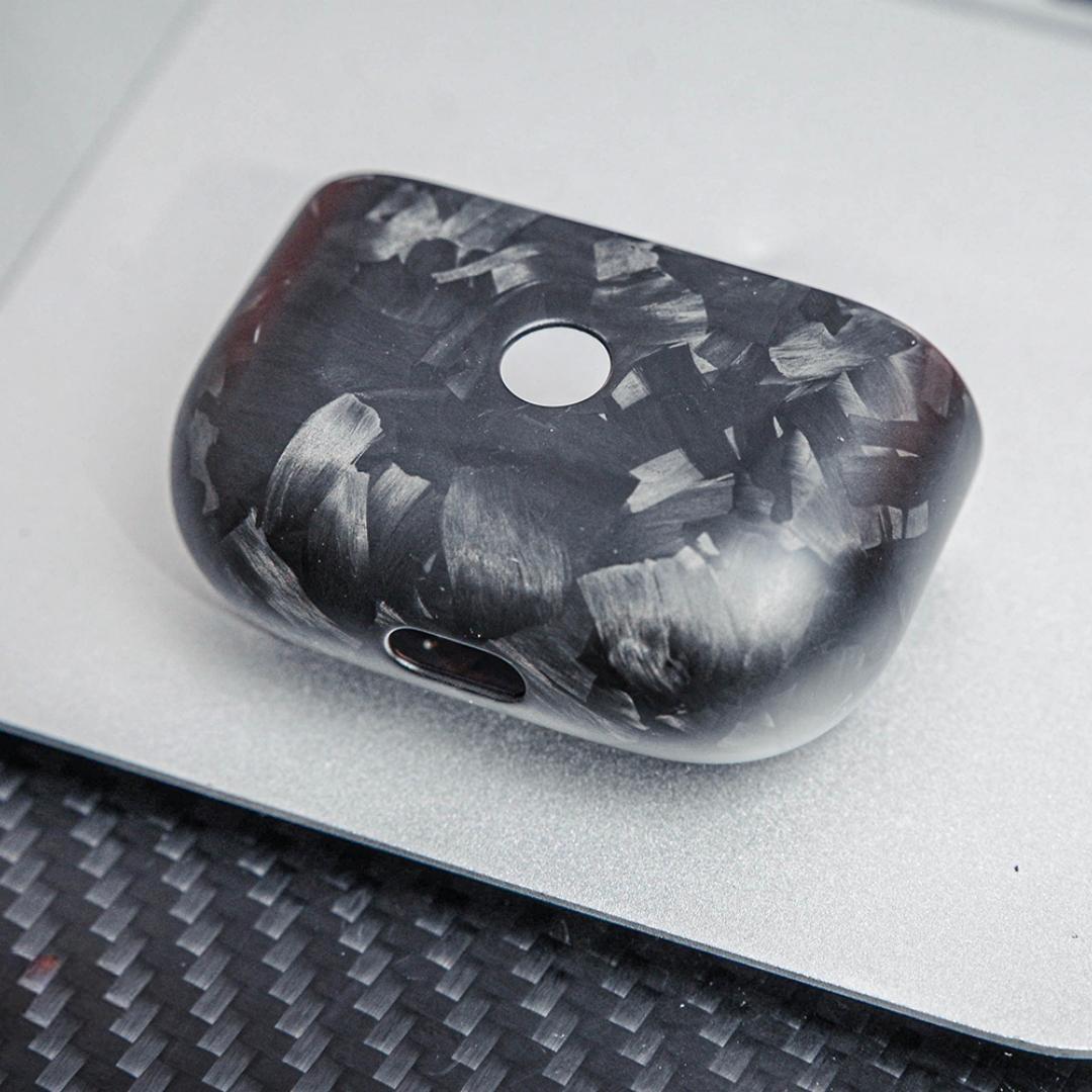 Close-up of the front side of the lower half of a forged carbon fiber AirPods case, showcasing its unique marbled texture, placed on a silver surface with a carbon fiber textured background.