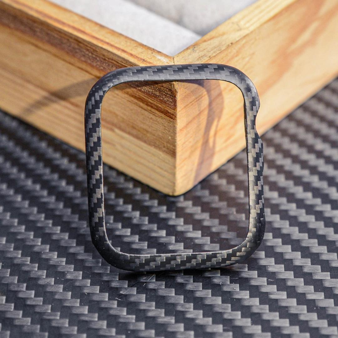 Close-up of a 3K carbon fiber Apple Watch case with a distinct woven texture, displayed on a carbon fiber-textured surface with wooden accents in the background.