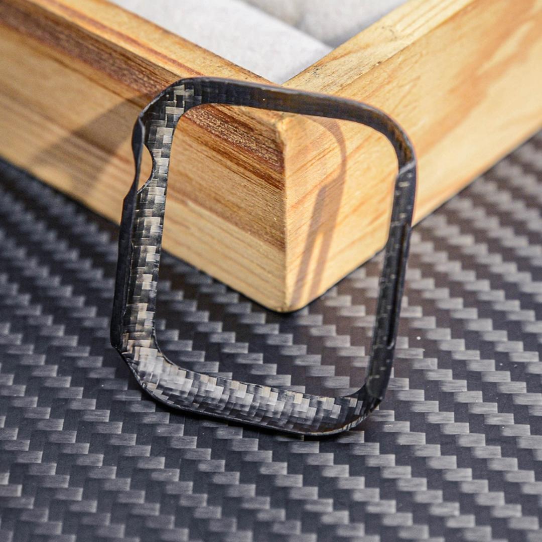 Close-up of a 3K carbon fiber Apple Watch case with a distinct woven texture, displayed on a carbon fiber-textured surface with wooden accents in the background.