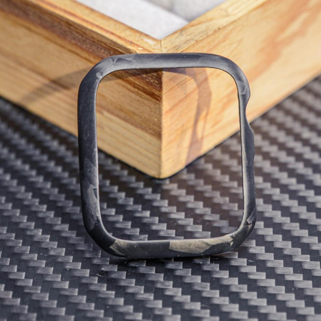 Close-up of a forged carbon fiber Apple Watch case with a distinct marbled texture, displayed on a carbon fiber-textured surface with wooden accents in the background.