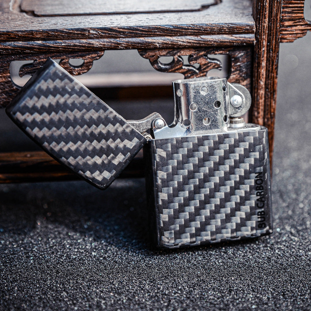 Close-up of an open Zippo lighter with a 3K carbon fiber case showcasing the distinct weave pattern and "BUB CARBON" branding, positioned against a carved wooden structure and a glittery surface.