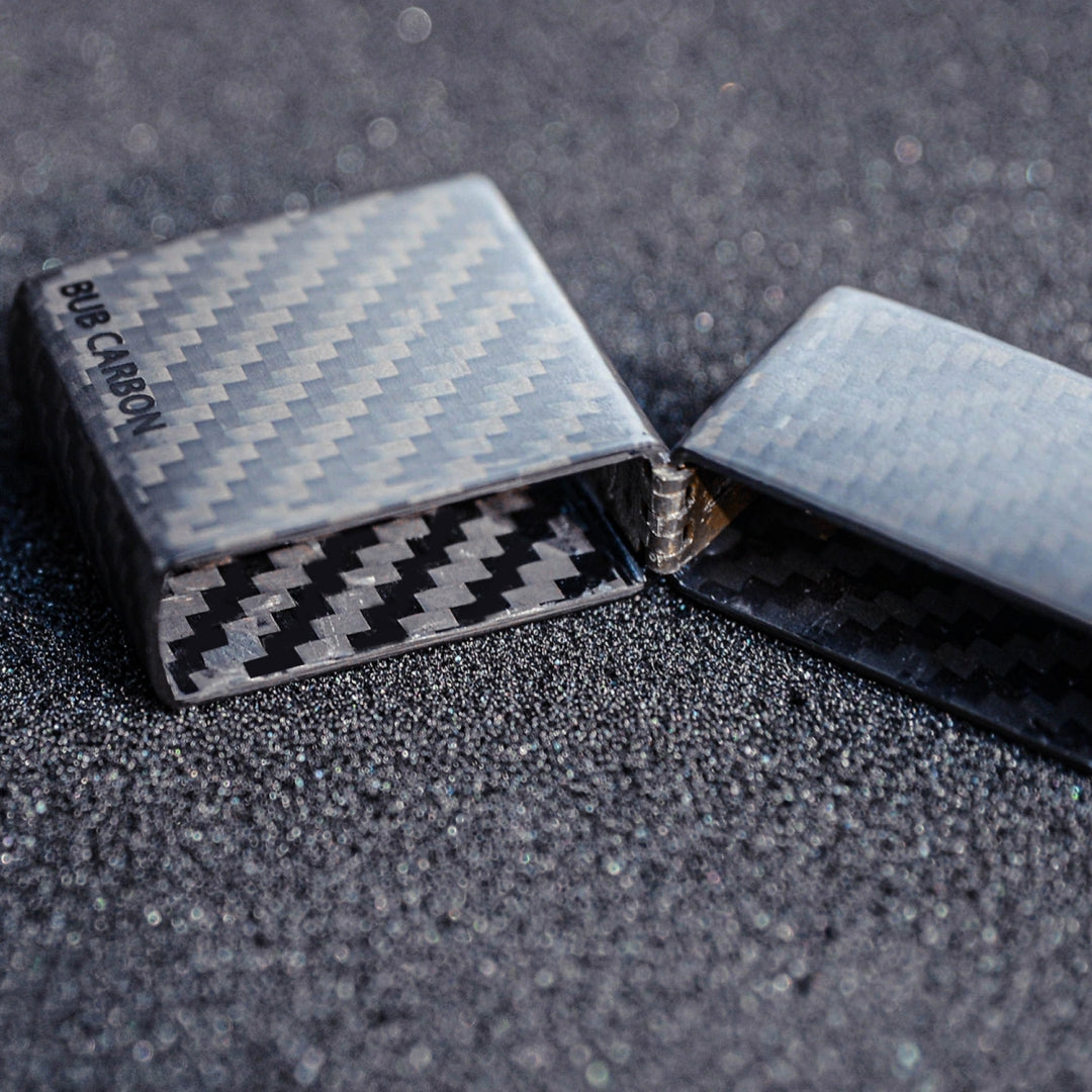 Close-up of an open 3K carbon fiber Zippo lighter case showcasing the detailed carbon fiber weave pattern both inside and out, with “BUB CARBON” branding, placed on a textured, glittery surface.