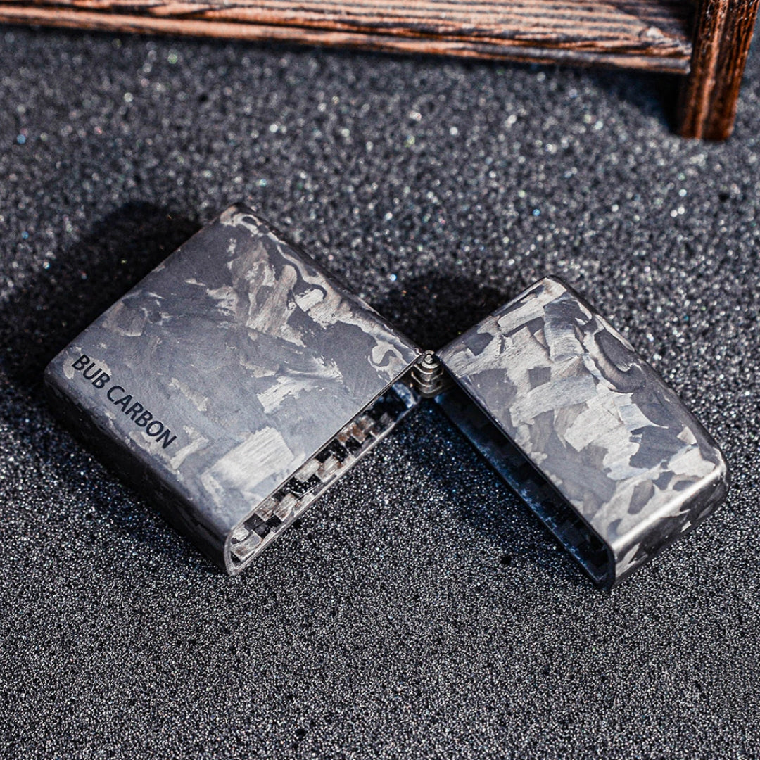 Close-up of an open forged carbon fiber Zippo lighter case with "BUB CARBON" branding, showcasing the unique marbled texture, placed on a textured dark glittery surface with a wooden structure in the background.