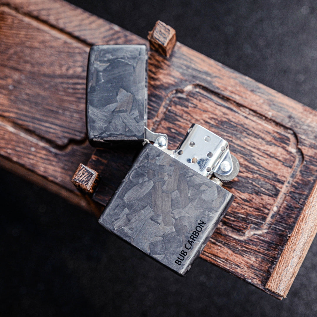 Close-up of an open Zippo lighter with a forged carbon fiber case featuring "BUB CARBON" branding, displayed on a textured wooden surface.