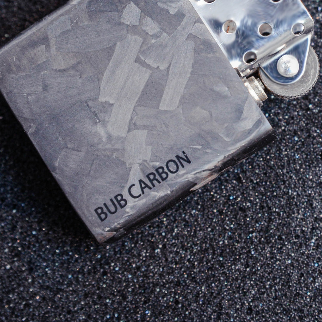 Close-up of a Zippo lighter case with forged carbon fiber construction, showcasing the detailed marbled texture and "BUB CARBON" branding