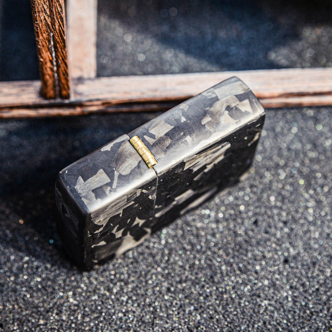 Close-up of a closed Zippo lighter case with forged carbon fiber construction, showcasing its unique marbled texture and brass hinge, placed on a textured dark glittery surface with a wooden structure in the background.