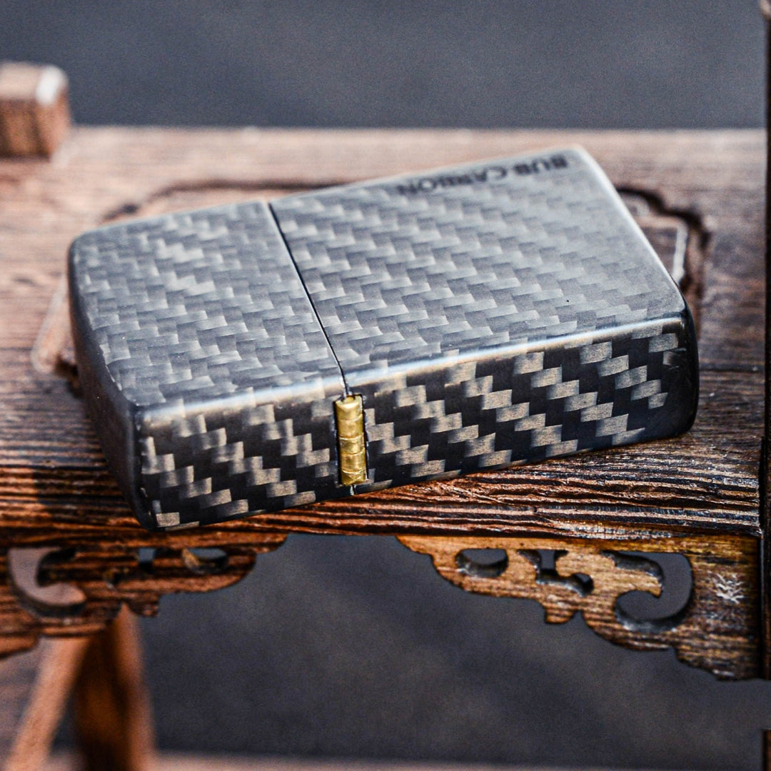 Close-up of a closed 3K carbon fiber Zippo lighter case showcasing the distinct weave pattern, brass hinge, and "BUB CARBON" branding, placed on a textured wooden surface with carved details.