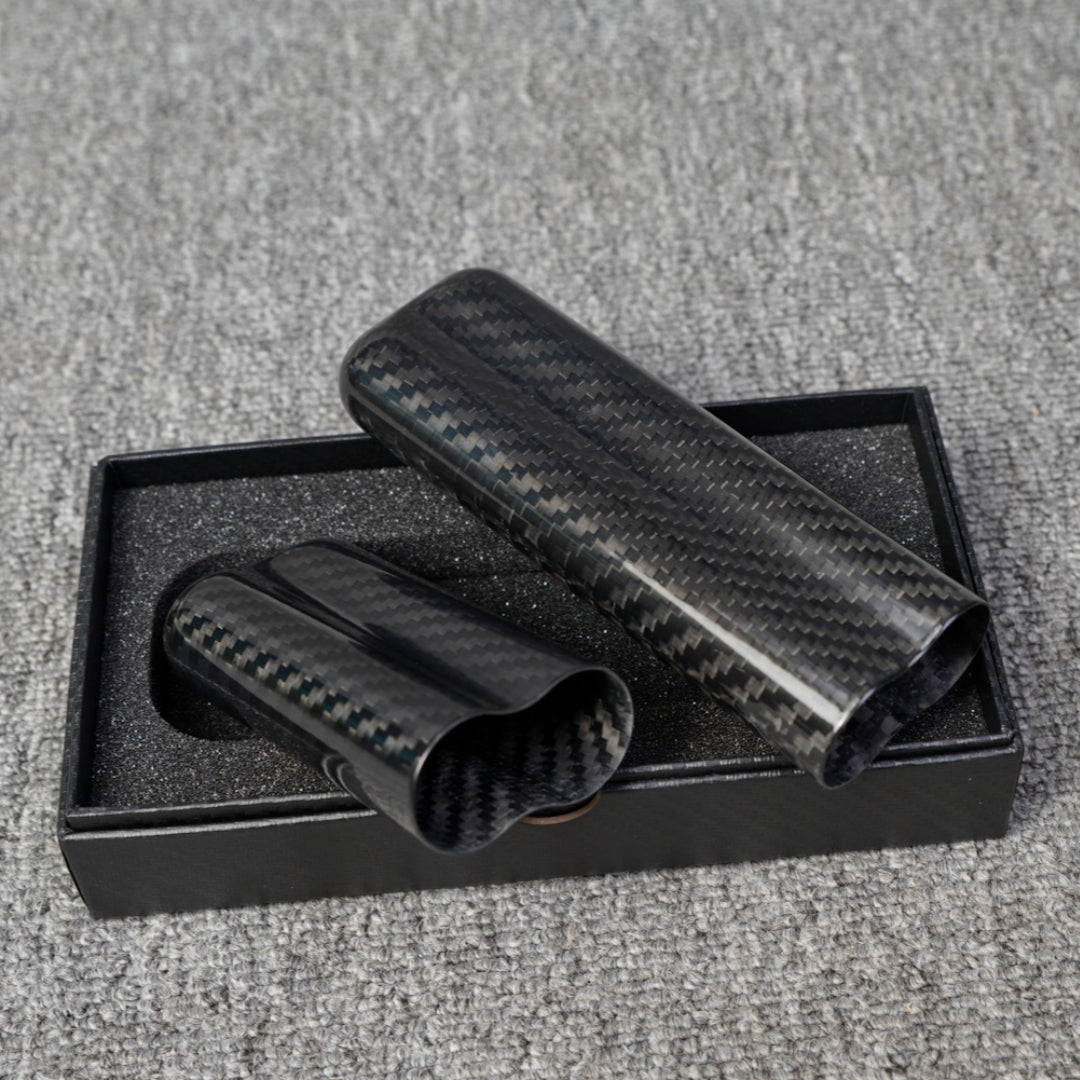 Close-up of two carbon fiber cigar tubes (capable of holding 2 cigars) with a distinct weave pattern, placed in a black box with foam lining on a gray fabric surface.