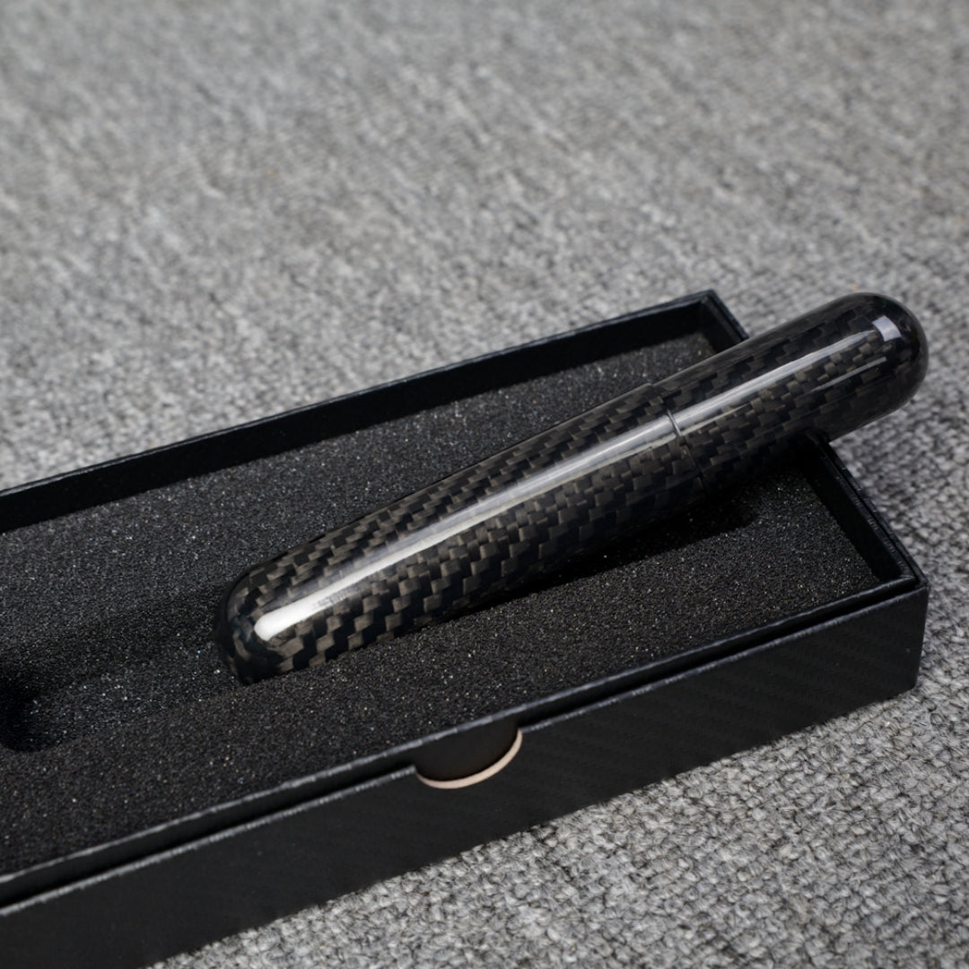 Close-up of a carbon fiber cigar tube (single cigar capacity) with a distinct weave pattern, placed in a black box with foam lining on a gray fabric surface.