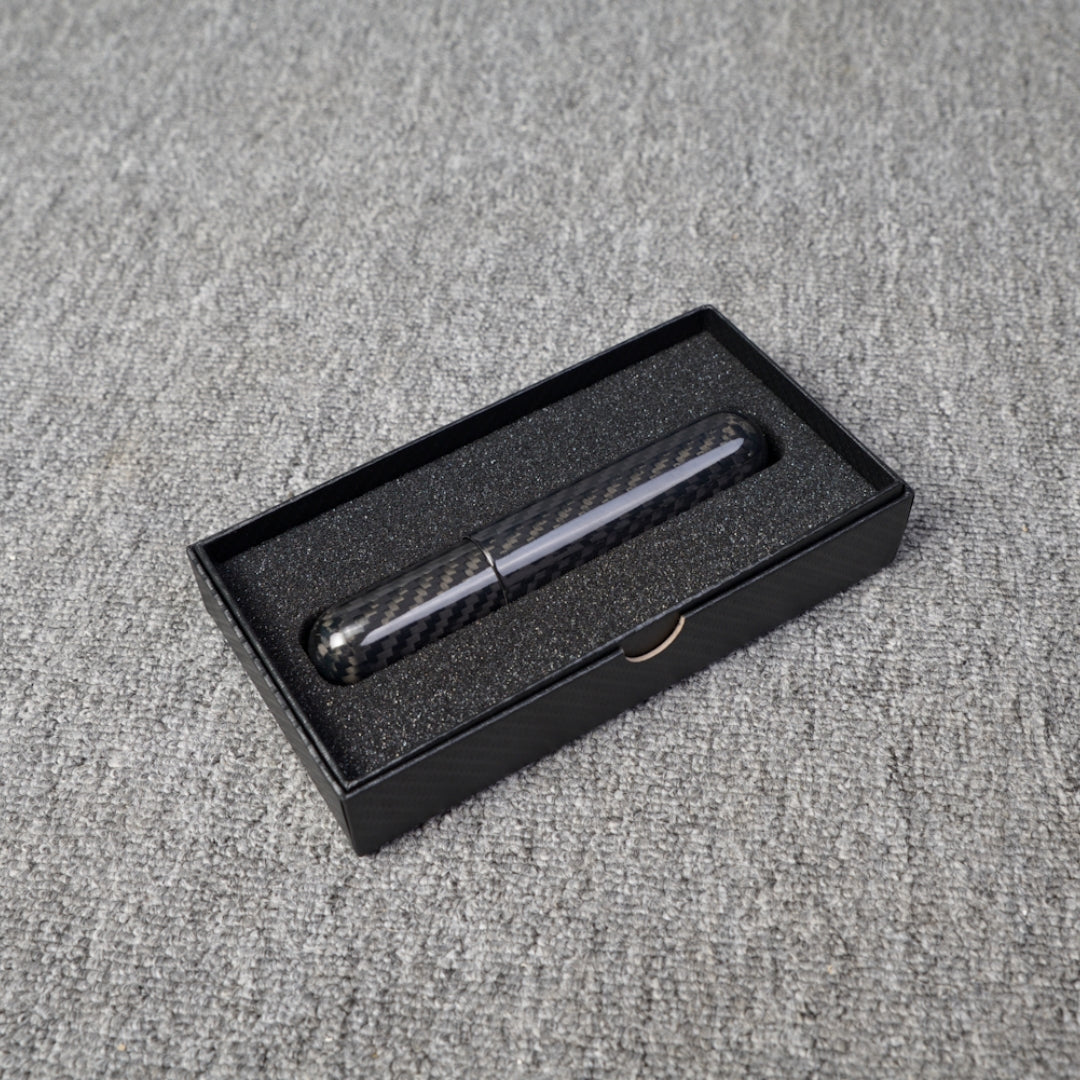 Close-up of a carbon fiber cigar tube with single cigar capacity, featuring a distinct weave pattern, placed in a black box with foam lining on a gray fabric surface.