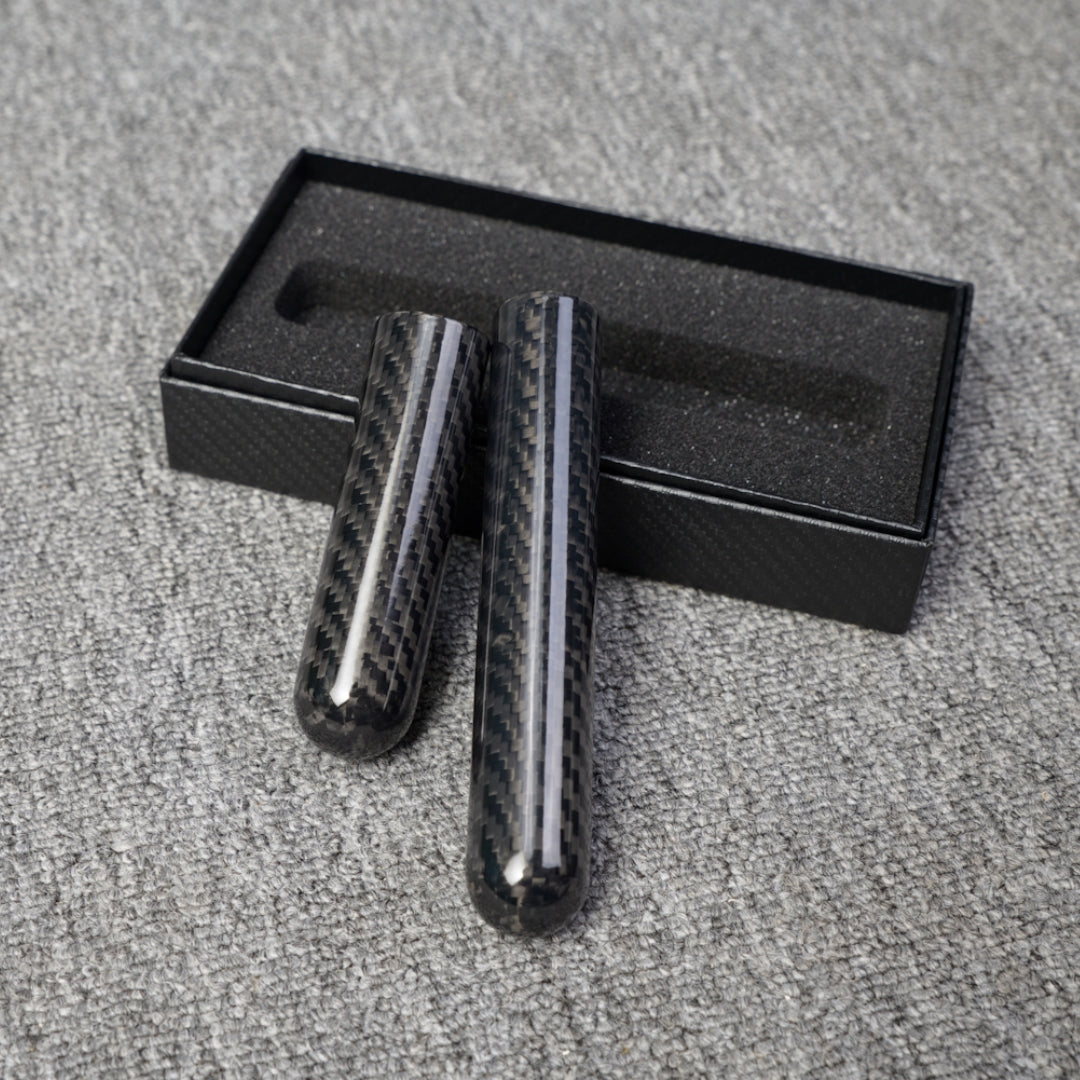 Close-up of two carbon fiber cigar tubes with single-cigar capacity, featuring a distinct woven pattern, placed next to an open black box with foam lining on a gray fabric surface.