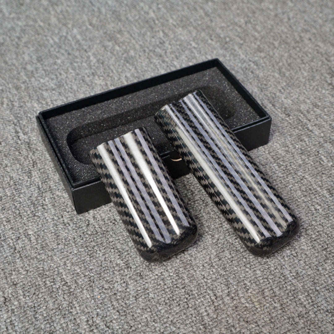 Close-up of two carbon fiber cigar tubes with a distinct striped carbon fiber pattern, placed near an open black box with foam lining on a gray fabric surface.