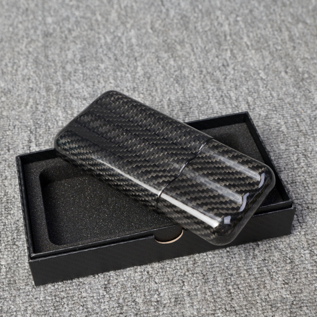 Close-up of a carbon fiber cigar tube with a capacity for 3 cigars, featuring a distinct woven pattern, placed on an open black box with foam lining over a gray fabric surface.