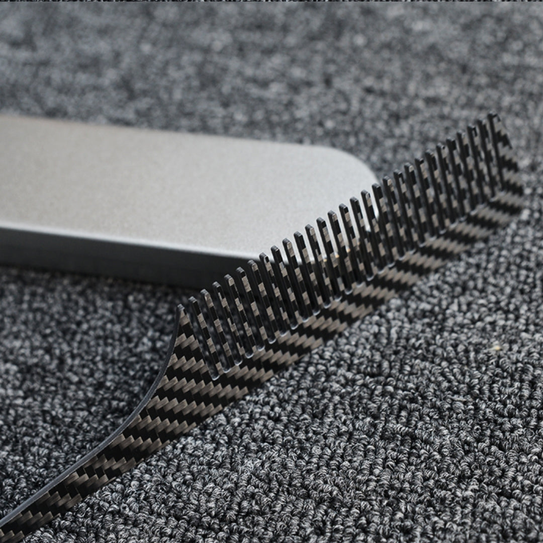 Close-up of a carbon fiber comb with a distinct woven pattern, placed next to a silver metal case on a dark gray textured fabric surface.