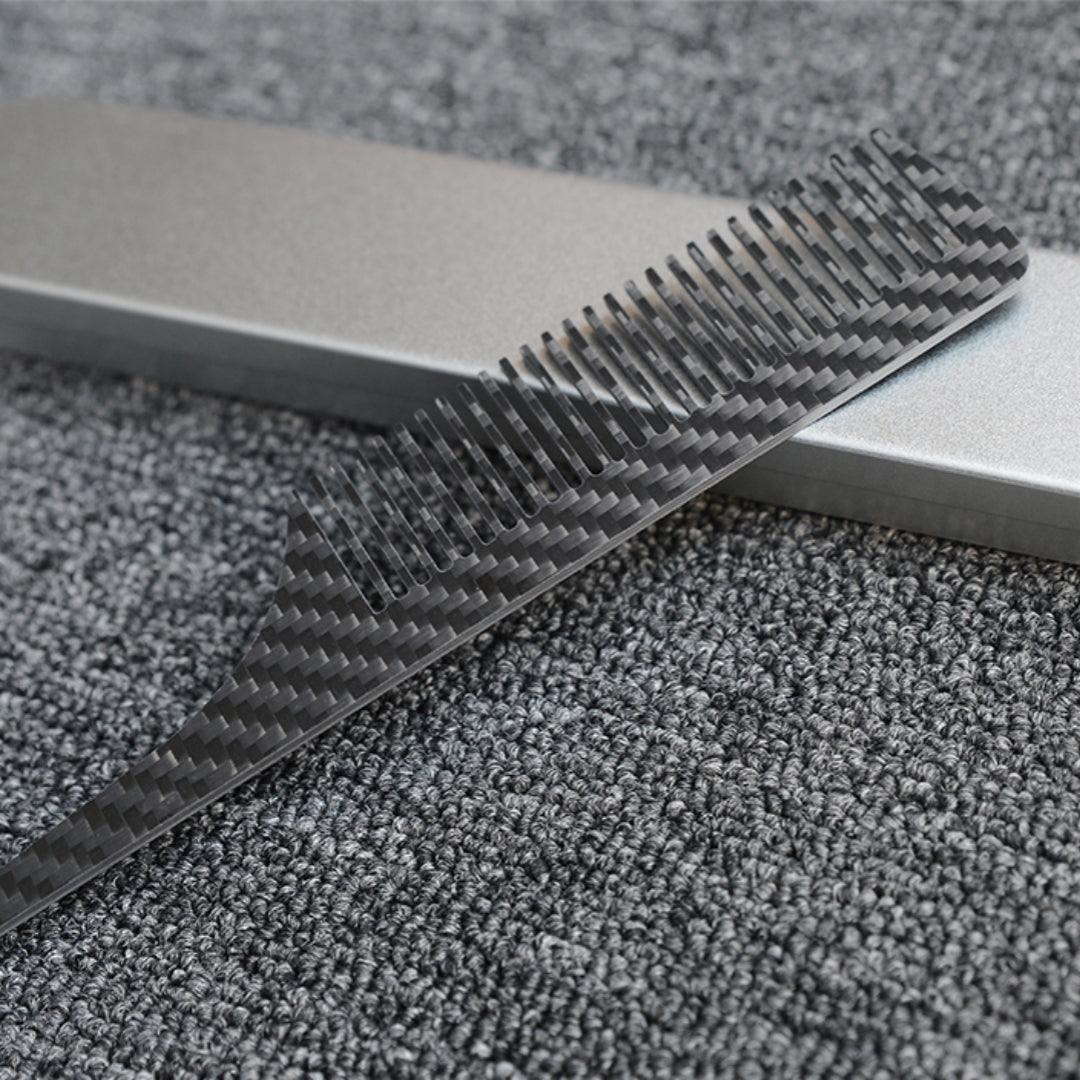 Close-up of a carbon fiber comb with a distinct woven pattern, placed next to a silver metal case on a dark gray textured fabric surface.
