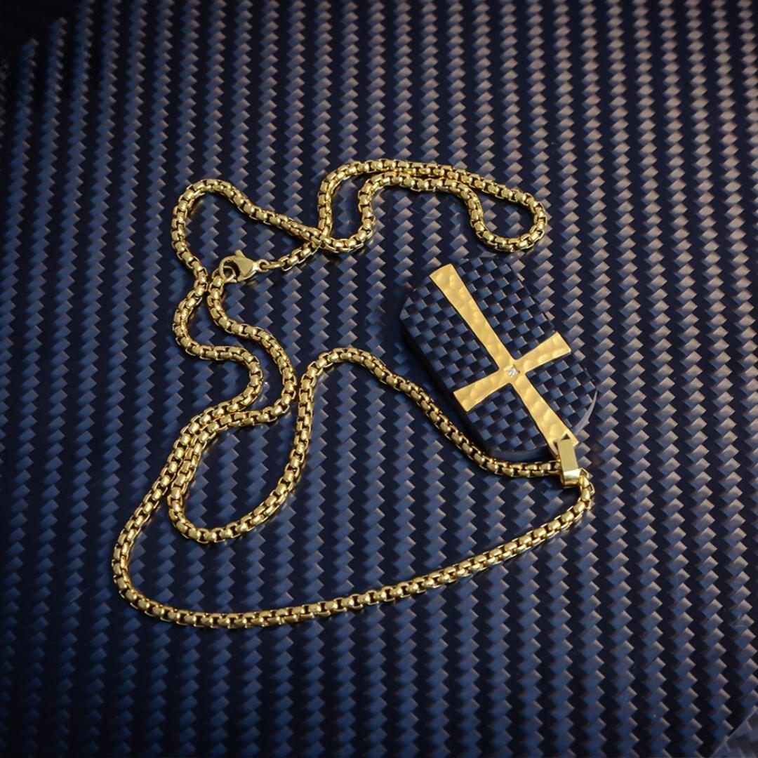 Close-up of a 3K carbon fiber pendant featuring a gold cross accent with a small diamond detail, paired with a gold-tone chain, displayed on a carbon fiber textured background.